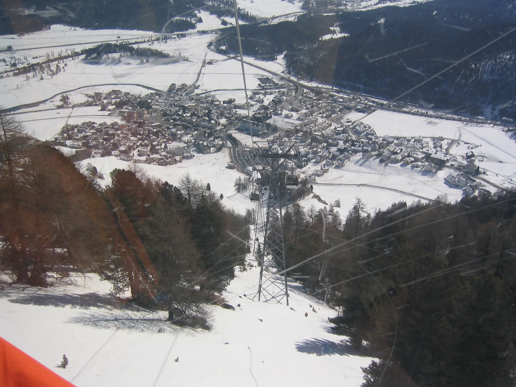A steep snowy slope and lift line lead toward buildings spread across a winter valley.