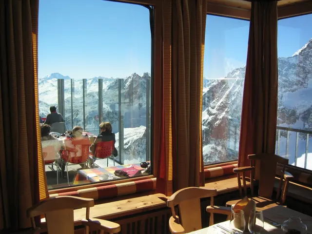 Wood-framed windows reveal red terrace chairs and snowy mountains beyond.