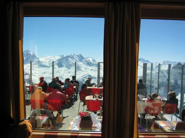 People sit in red chairs on a mountain terrace seen through tall windows.