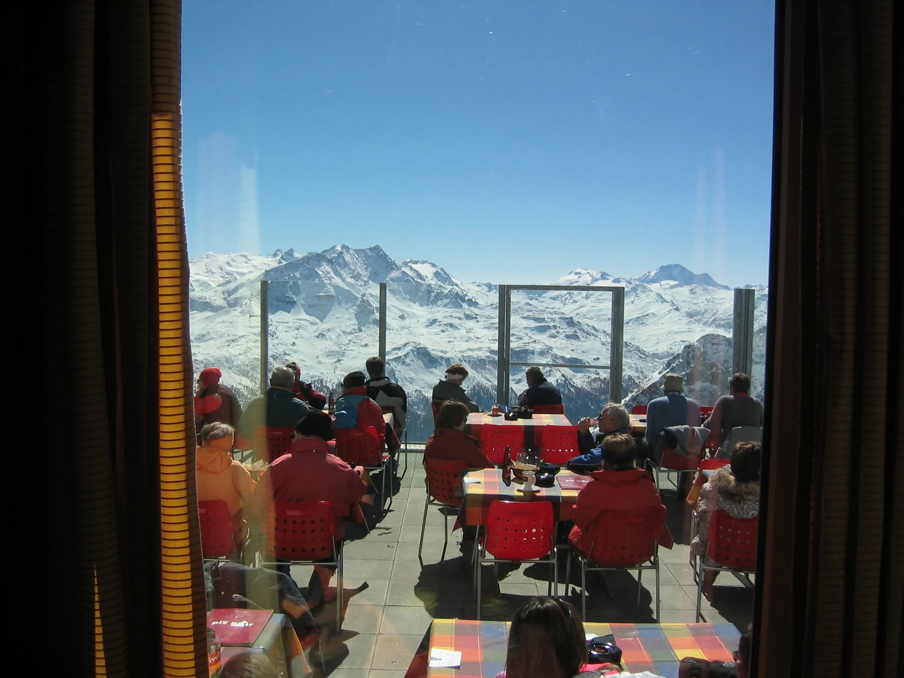 A window frames visitors on a terrace with red chairs and snow-covered peaks.