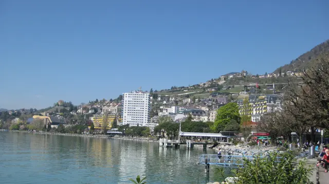 Waterfront buildings and hillside houses reflect in calm blue lake water.