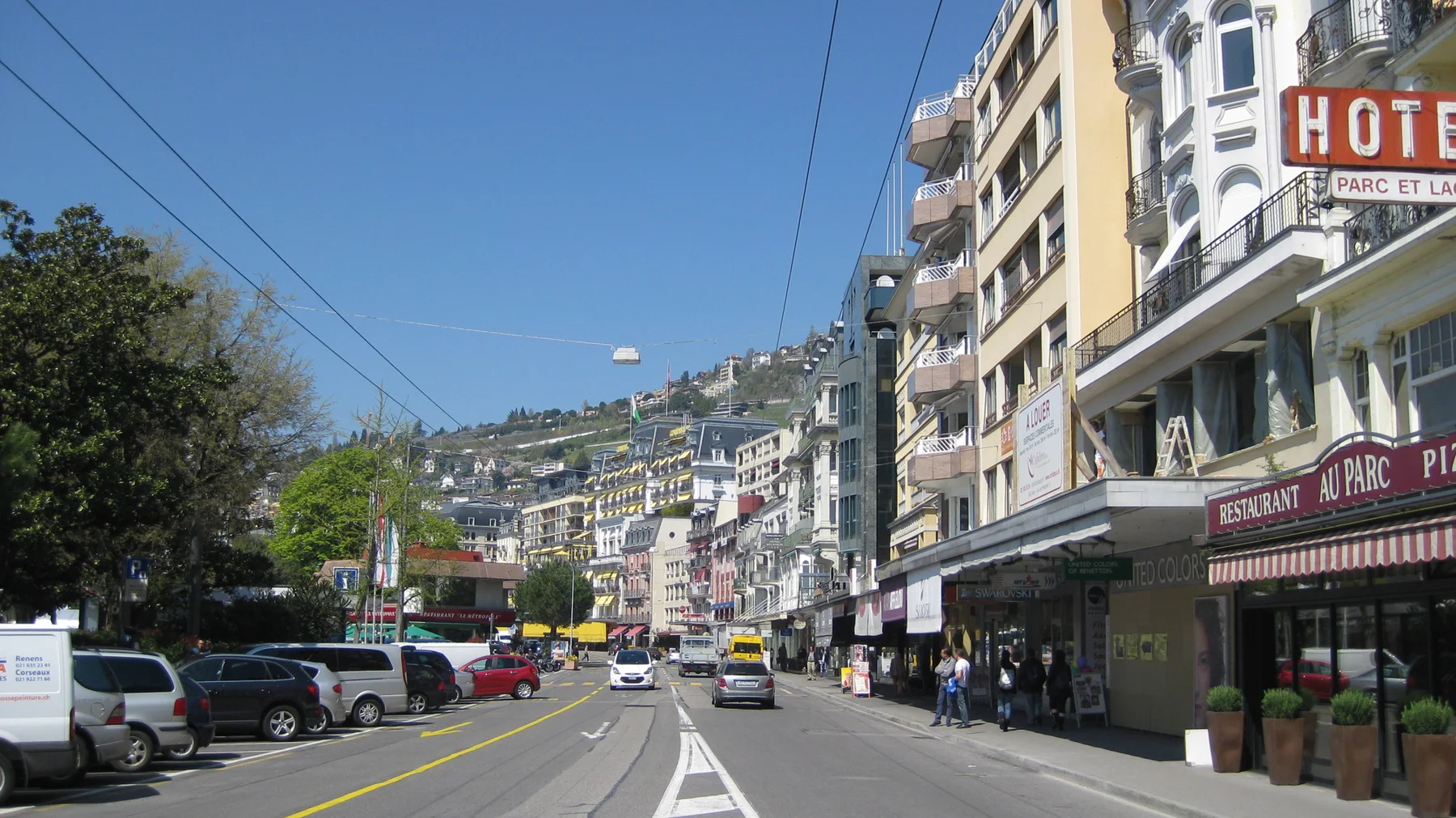 Cars travel past shops and tall buildings below a hillside covered with homes.