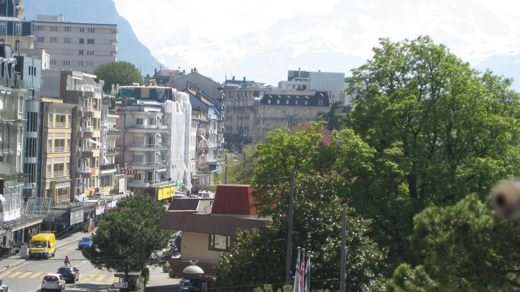Pale multistory buildings rise above a street and leafy trees near the lake.