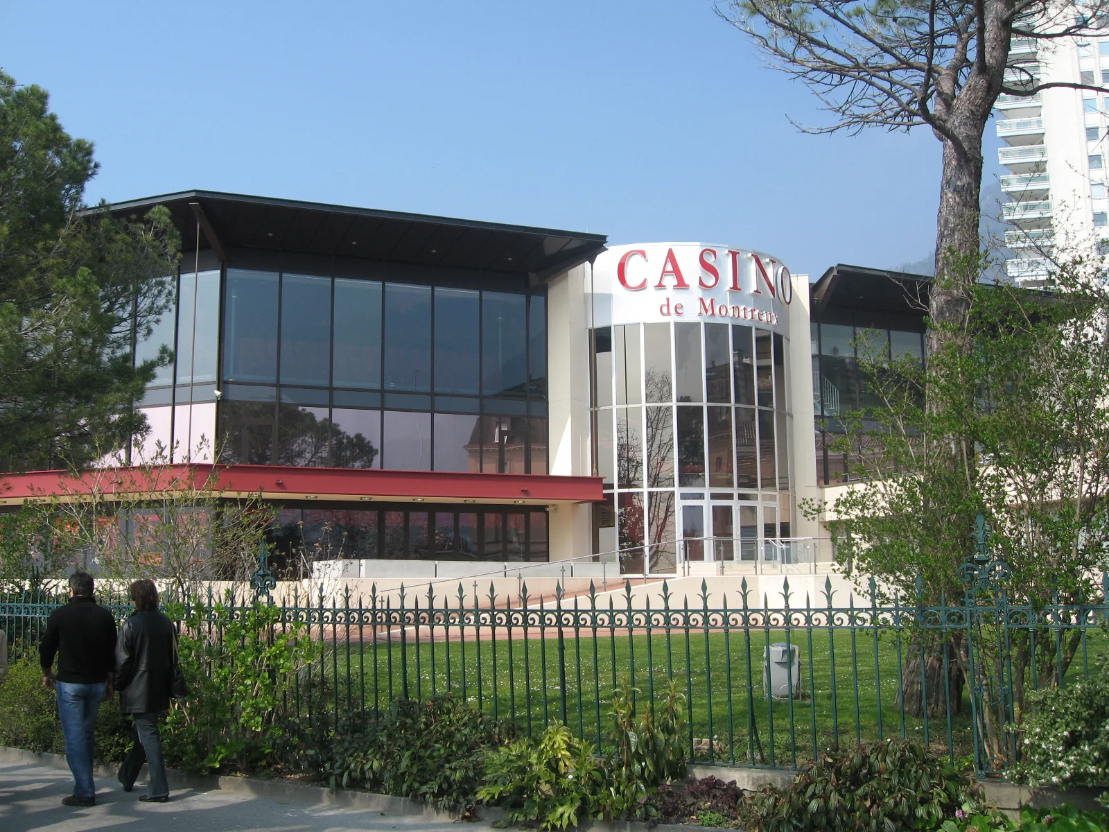 A glass-fronted casino building stands behind a garden and iron fence.