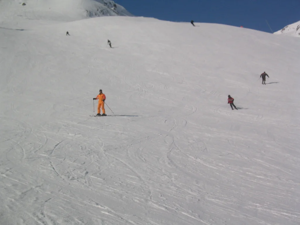 A skier in orange stands out among several figures on a wide snowy slope.