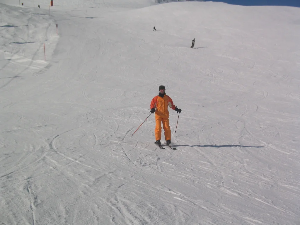 A skier dressed in orange pauses with poles on a tracked snow slope.