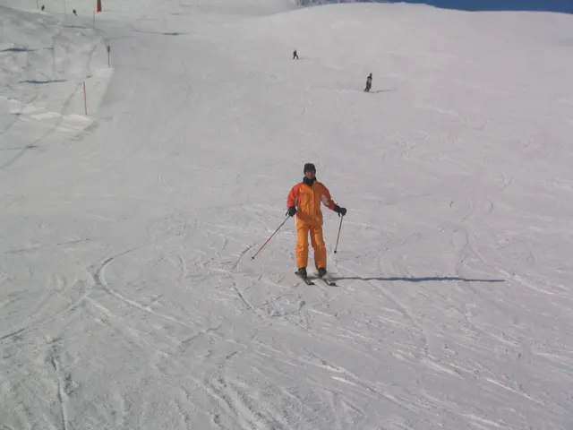 A skier dressed in orange pauses with poles on a tracked snow slope.