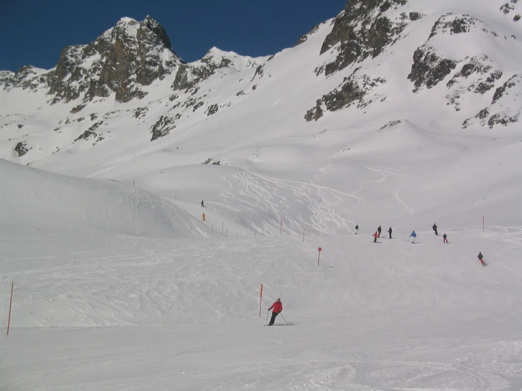 Small skiers form a loose line across a wide slope beneath rocky peaks.