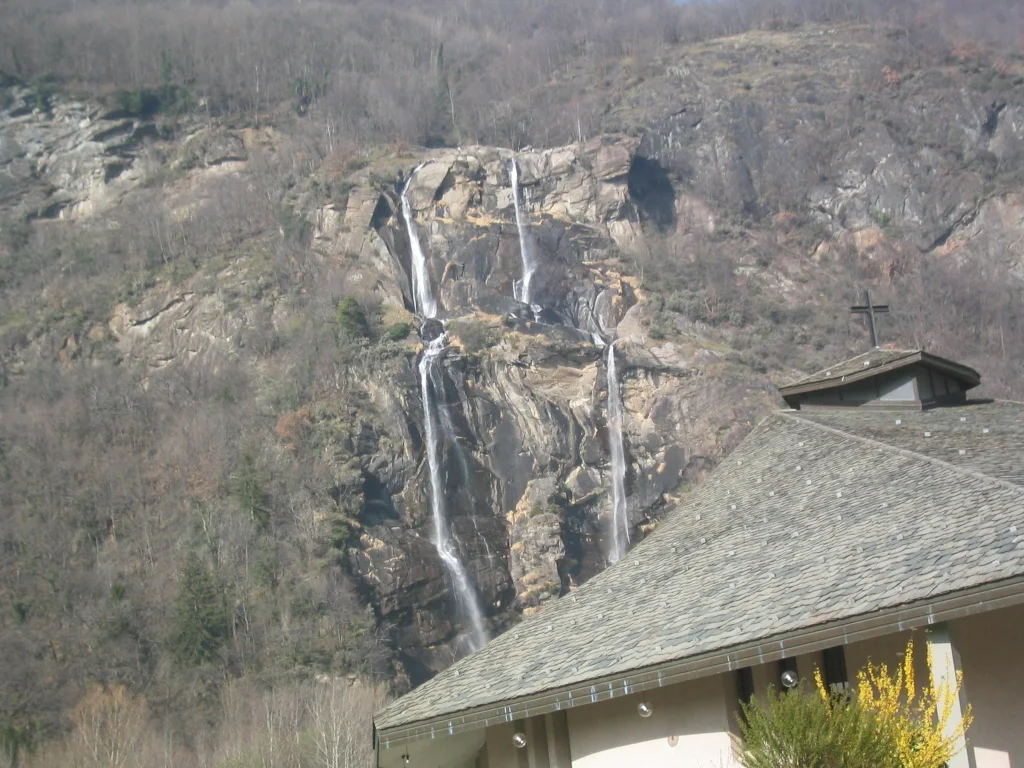 A narrow waterfall drops down a rocky mountainside behind a gray roof.