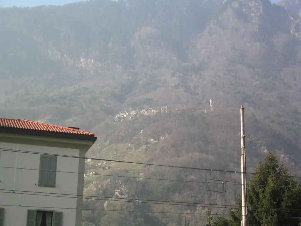 A pale building and roof edge sit beneath a hazy wooded mountain slope.
