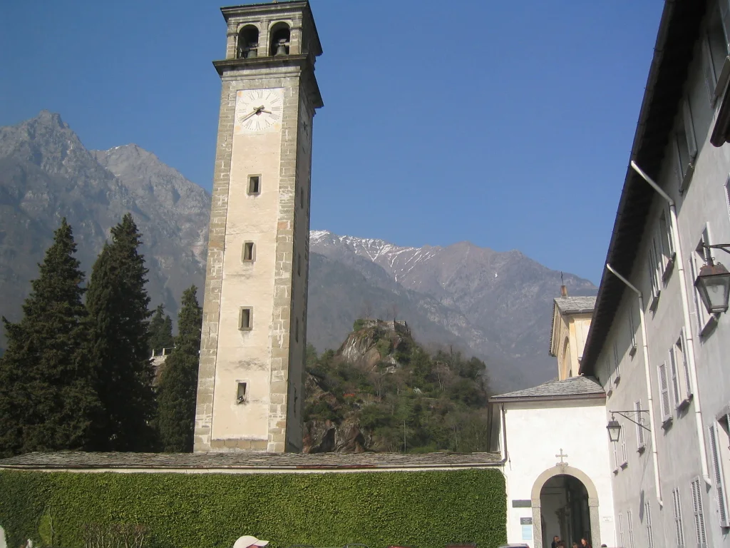 A slender stone clock tower rises beside a pale building with mountains behind it.