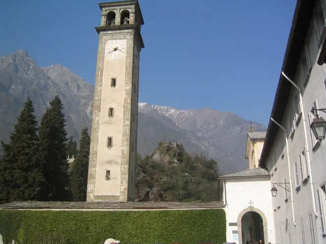 A slender stone clock tower rises beside a pale building with mountains behind it.