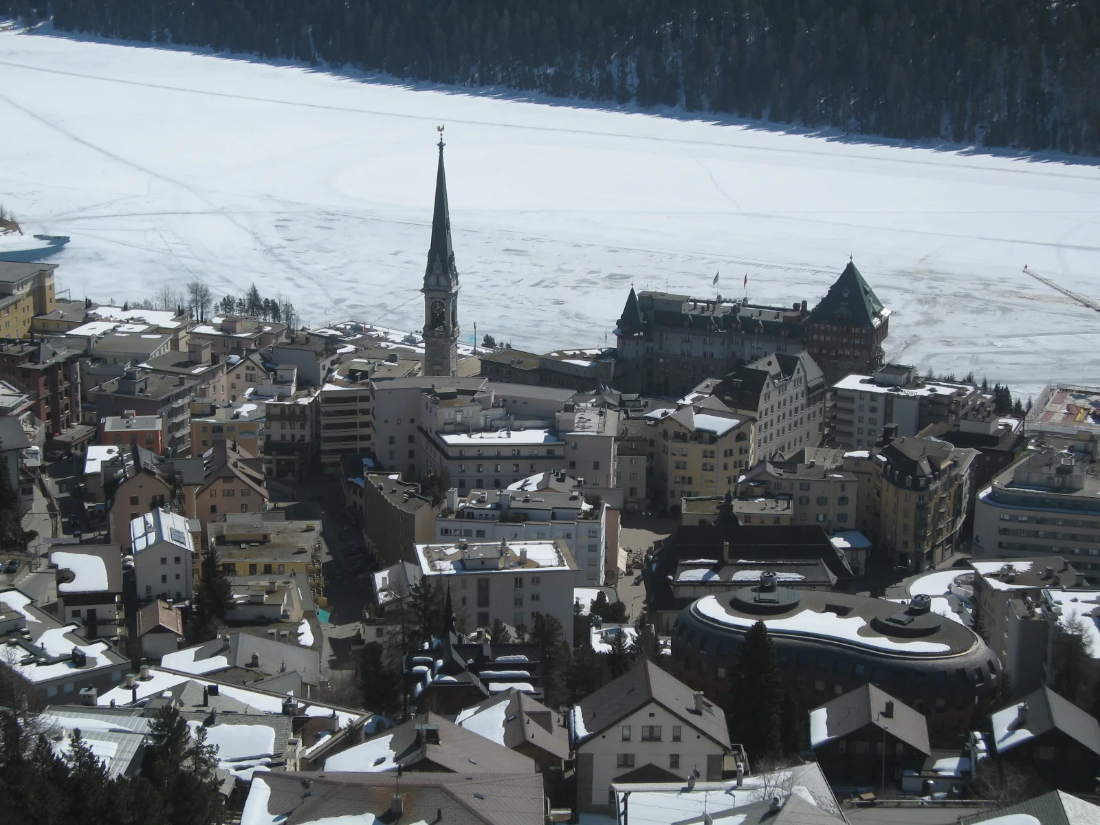 Snow-covered roofs and a slender church spire sit beside a frozen lake.