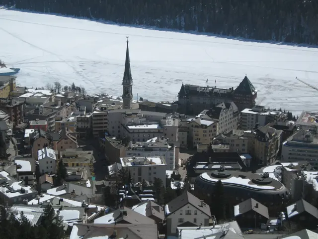 Snow-covered roofs and a slender church spire sit beside a frozen lake.
