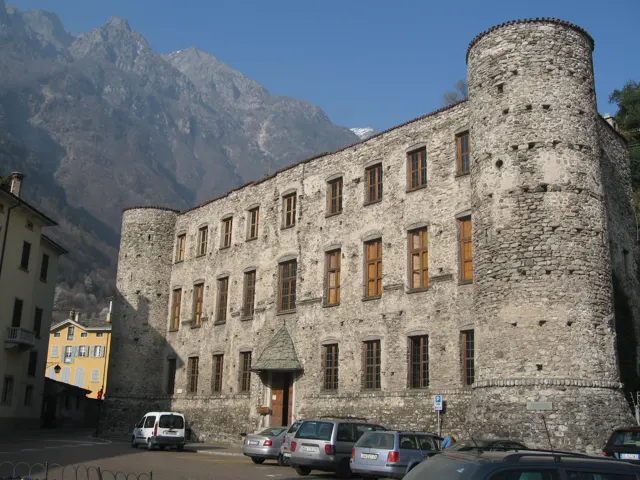 A long stone building with round corner towers stands beside a street below mountains.