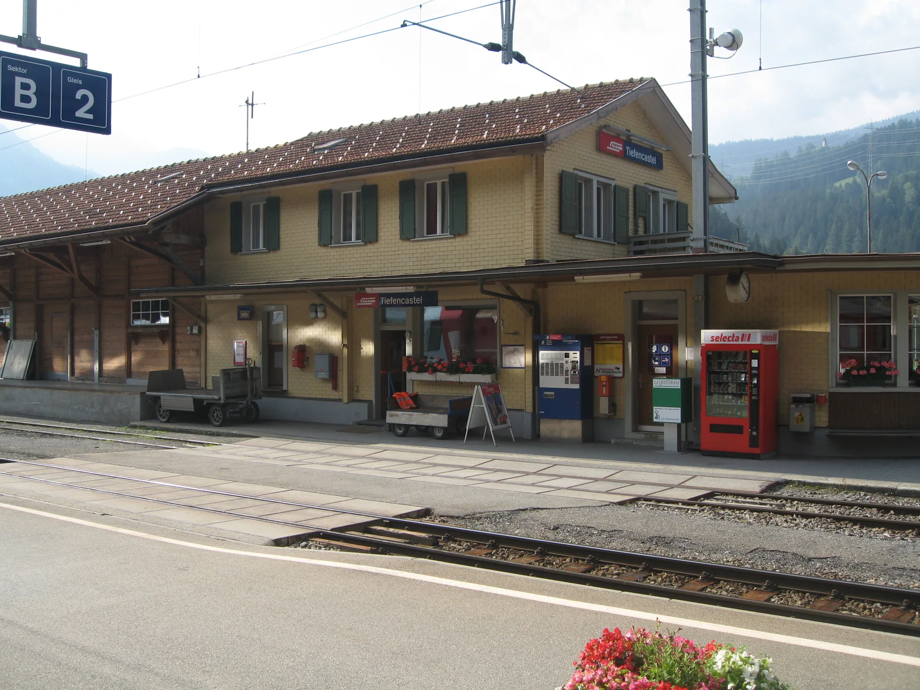 Railway tracks run past a small yellow station building and a platform sign.