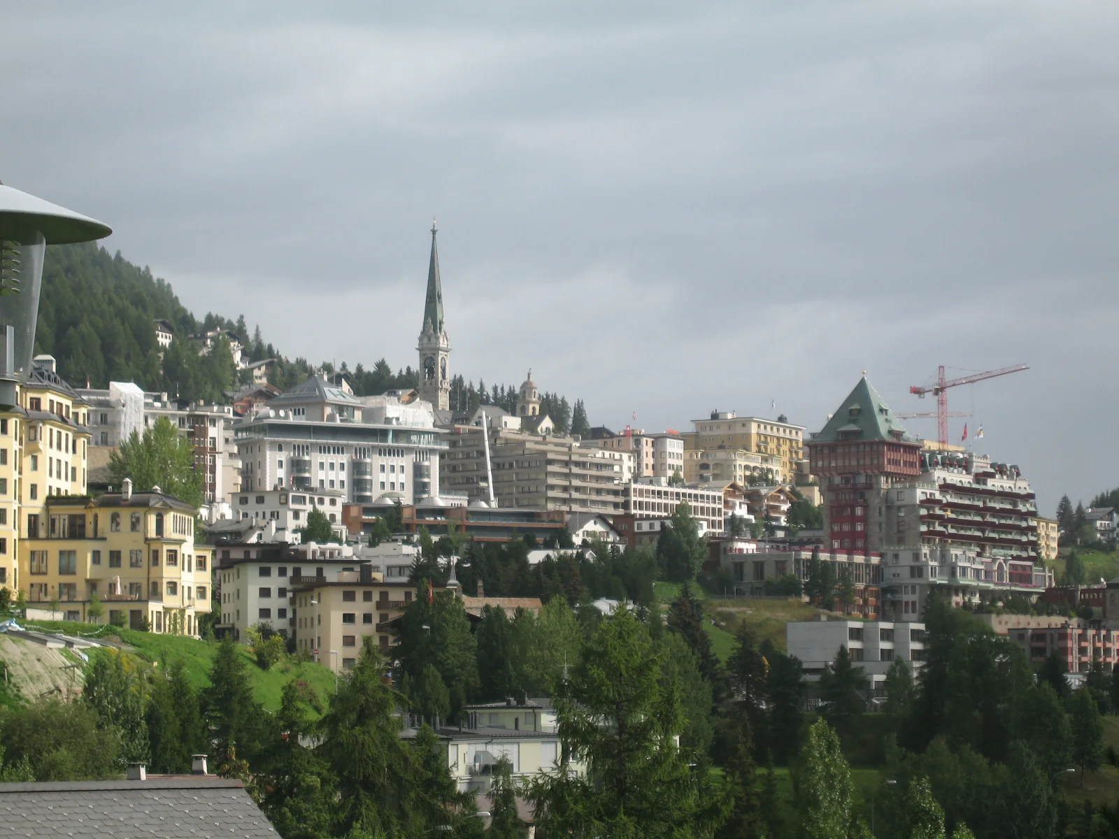 Tall buildings and a church spire cluster on a wooded hillside beneath gray clouds.