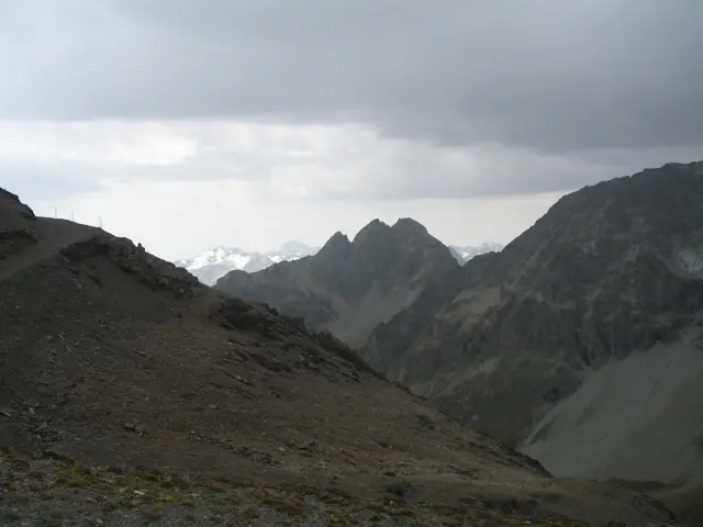 A stony mountainside leads toward distant jagged peaks under heavy cloud.