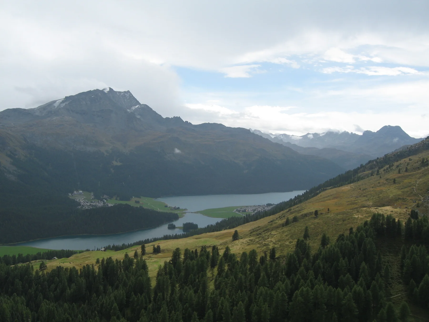 Clouds shade a mountain valley with lakes, dark forest and a golden-green hillside.