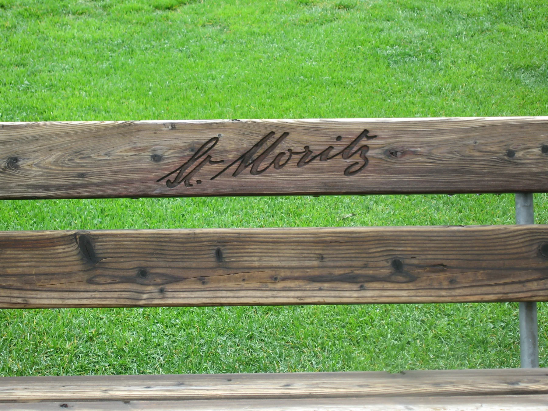 The words St. Moritz are carved into a wooden bench back above a green lawn.