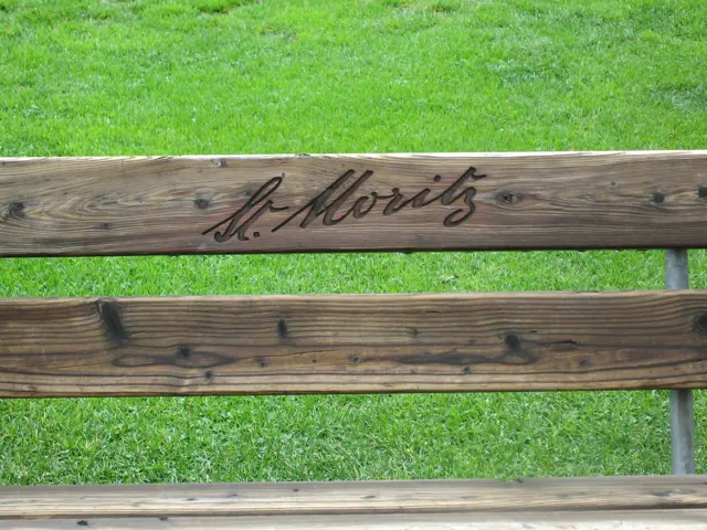 The words St. Moritz are carved into a wooden bench back above a green lawn.