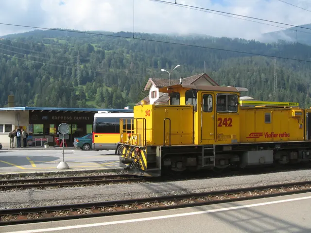 A yellow locomotive stands on station tracks beneath overhead wires and wooded hills.