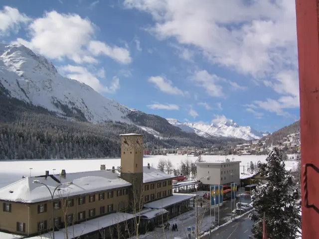 Snow-covered station buildings sit below a snowy mountain and a partly cloudy sky.