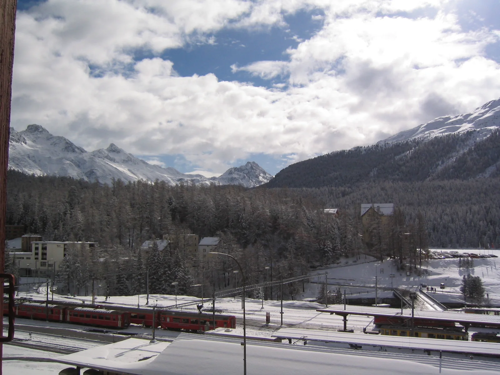 A red train stands along snowy tracks beneath forested slopes and white peaks.