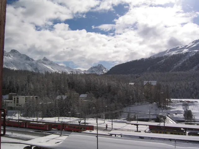A red train stands along snowy tracks beneath forested slopes and white peaks.