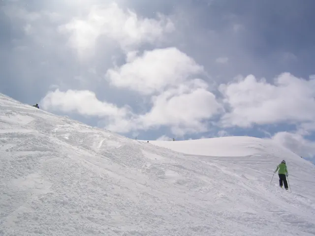A figure in a green jacket crosses a snowy slope under bright clouds.