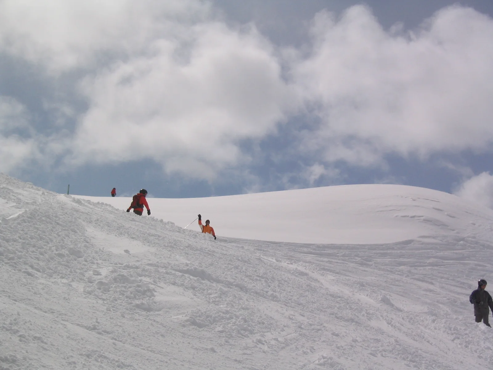 Several small figures stand near a snowy crest beneath a cloud-filled sky.