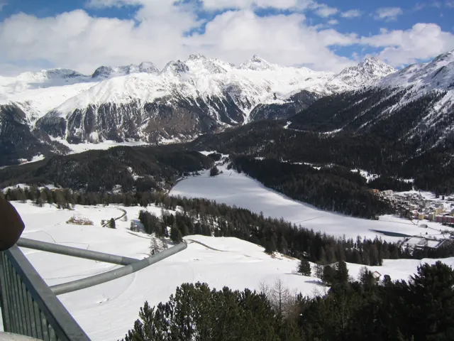 A frozen lake and dark forest fill a winter valley viewed from above a snowy slope.