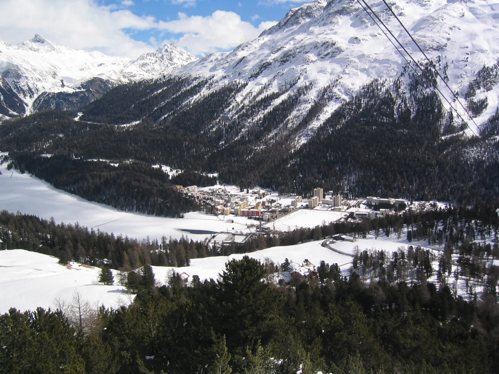 Lift cables cross a view of a frozen lake, trees and snowy mountains.