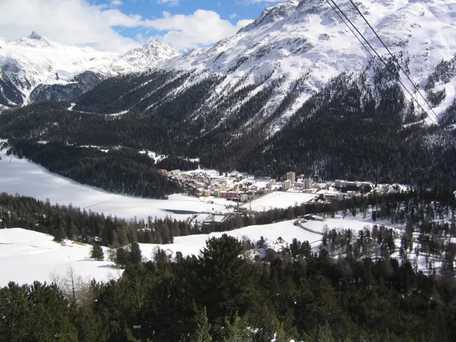 Lift cables cross a view of a frozen lake, trees and snowy mountains.