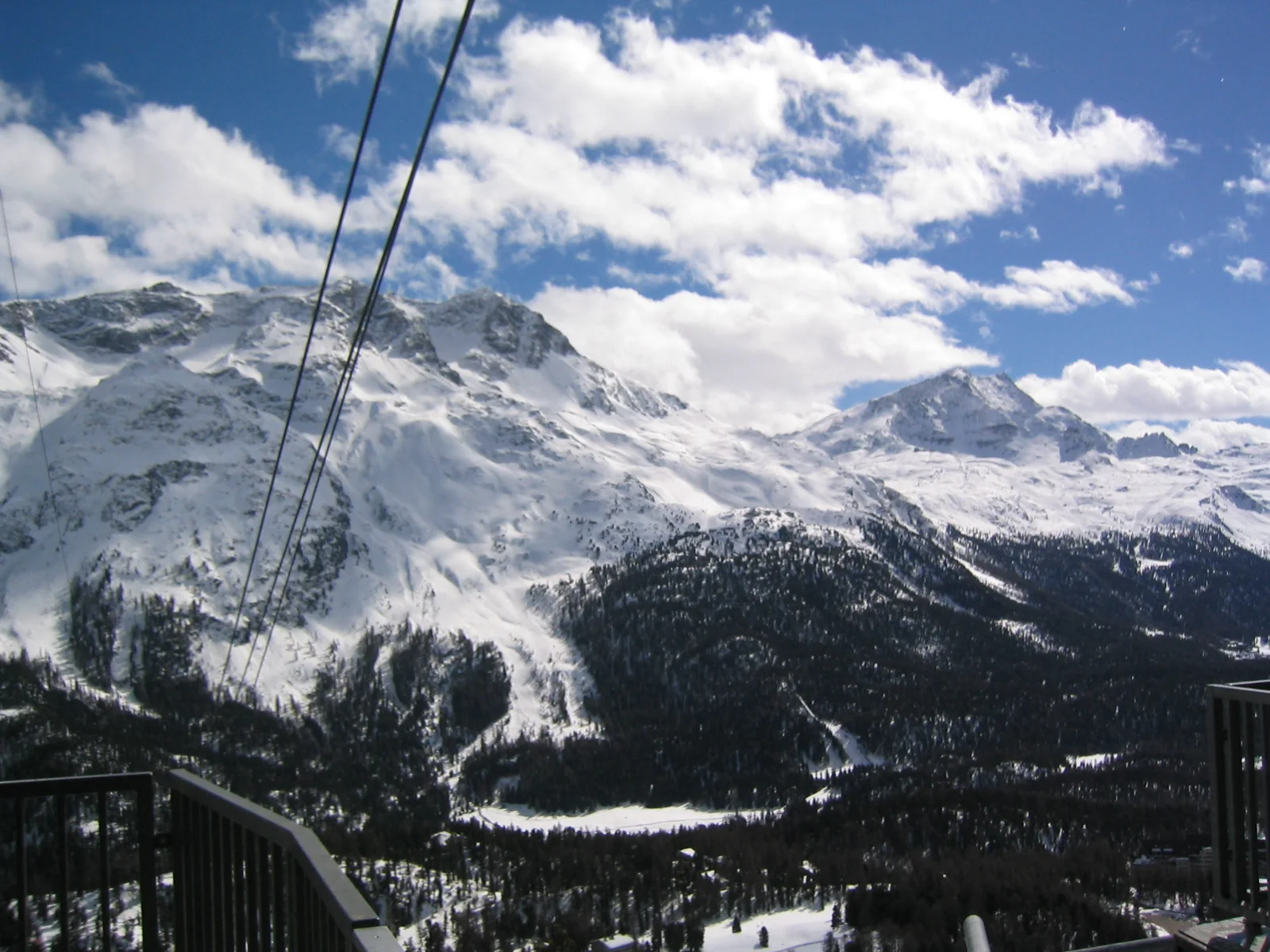 Lift cables lead toward snow-covered peaks beneath a bright, broken cloud cover.