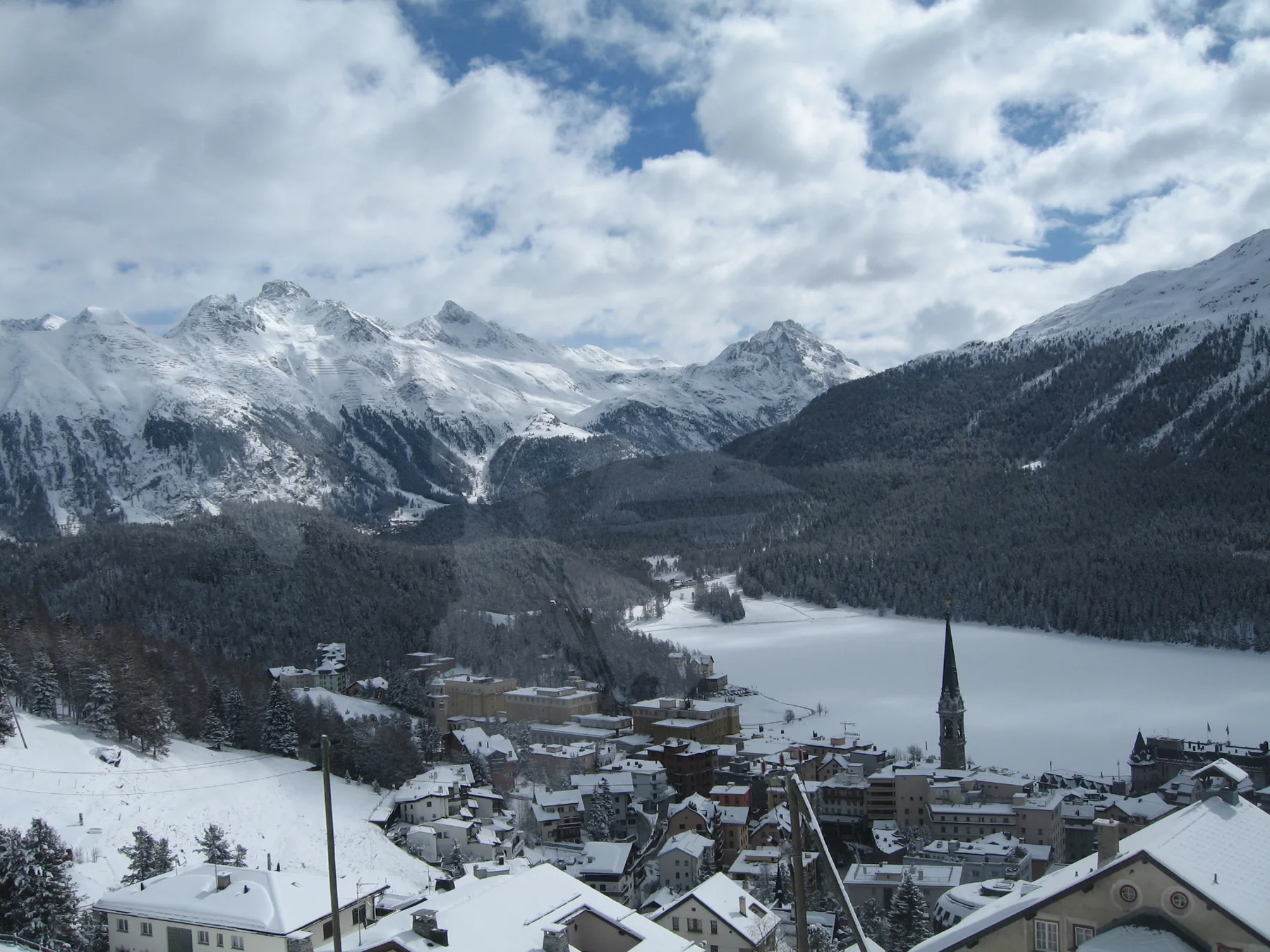 Snowy rooftops and a church spire overlook a frozen lake surrounded by mountains.