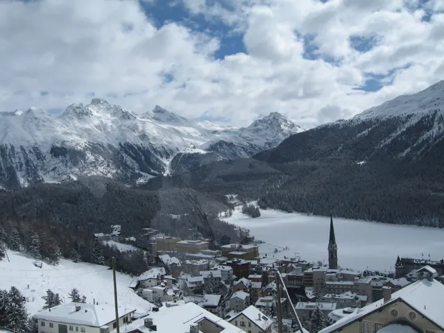 Snowy rooftops and a church spire overlook a frozen lake surrounded by mountains.