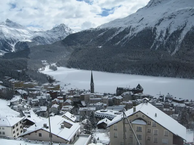 A slender spire rises among snow-covered buildings beside a frozen lake.