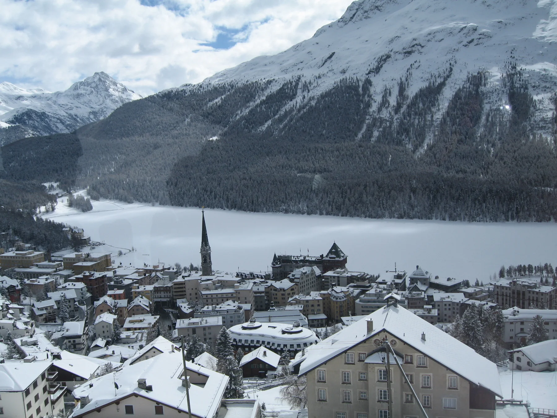A town spreads below a steep viewpoint beside a snow-covered lake and dark forest.