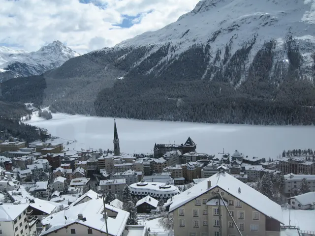A town spreads below a steep viewpoint beside a snow-covered lake and dark forest.