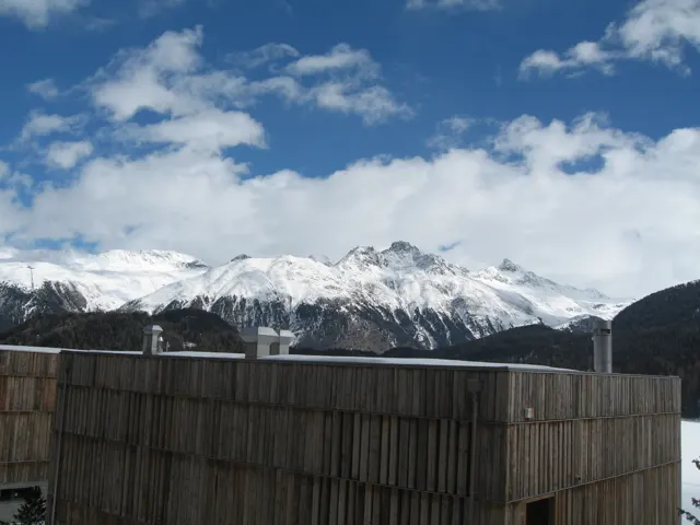 Snow-covered peaks rise beyond a tall wooden fence under a partly blue sky.