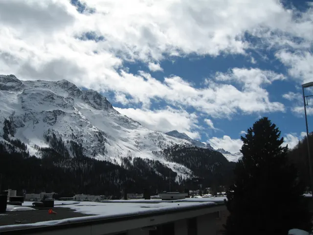 A low roof and dark tree frame bright snowy mountains and broken clouds.