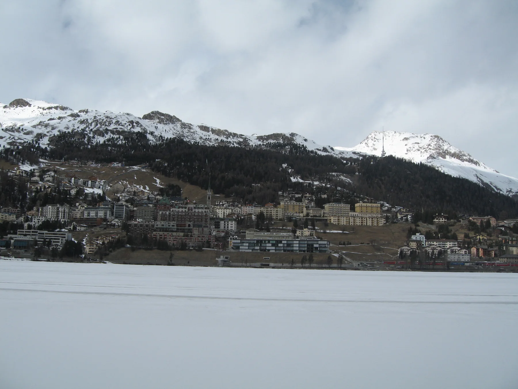 Dense hillside buildings overlook a flat snow-covered lake beneath snowy ridges.
