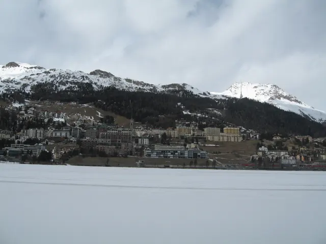 Dense hillside buildings overlook a flat snow-covered lake beneath snowy ridges.
