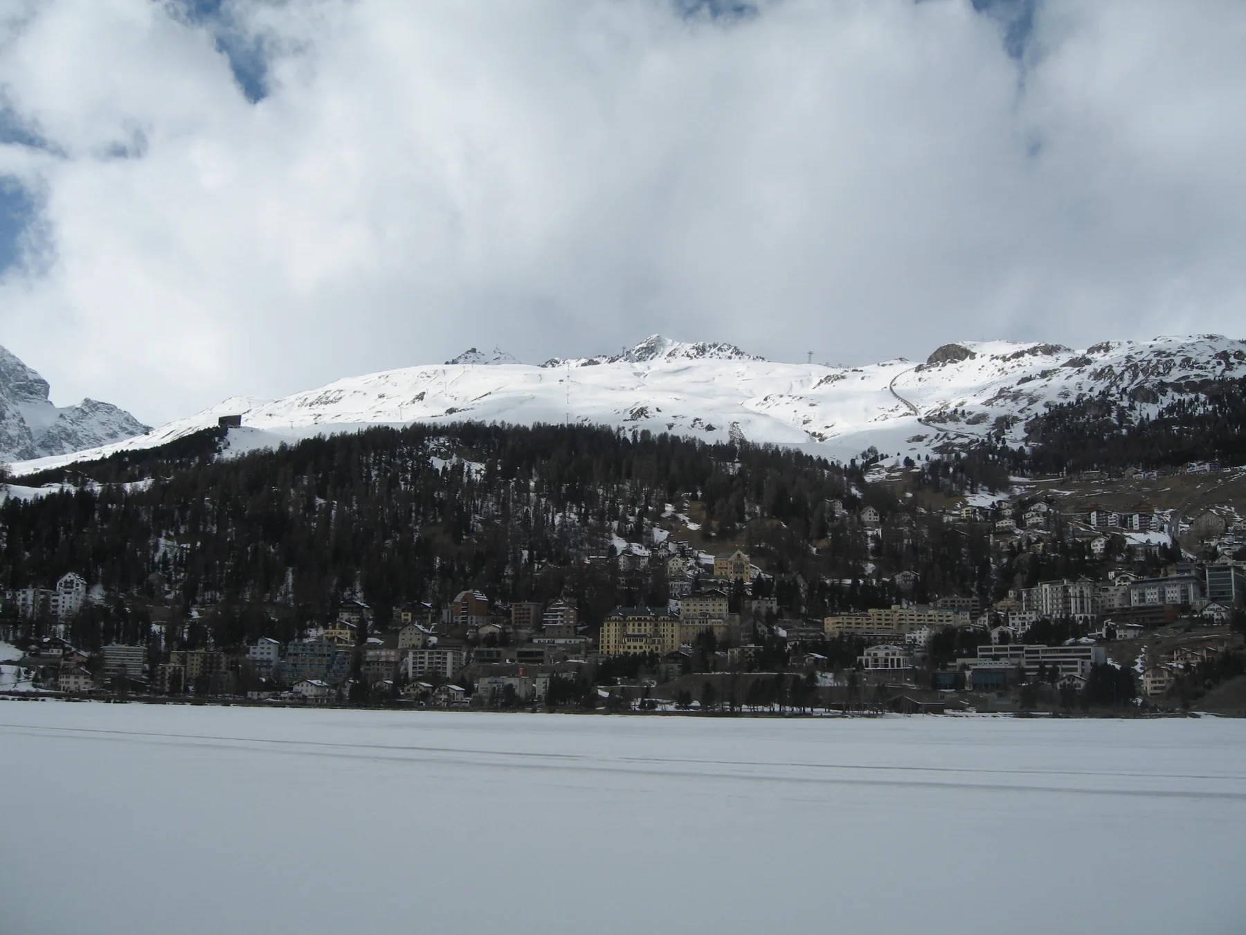 A broad frozen lake lies below wooded slopes and a snow-covered ridge.