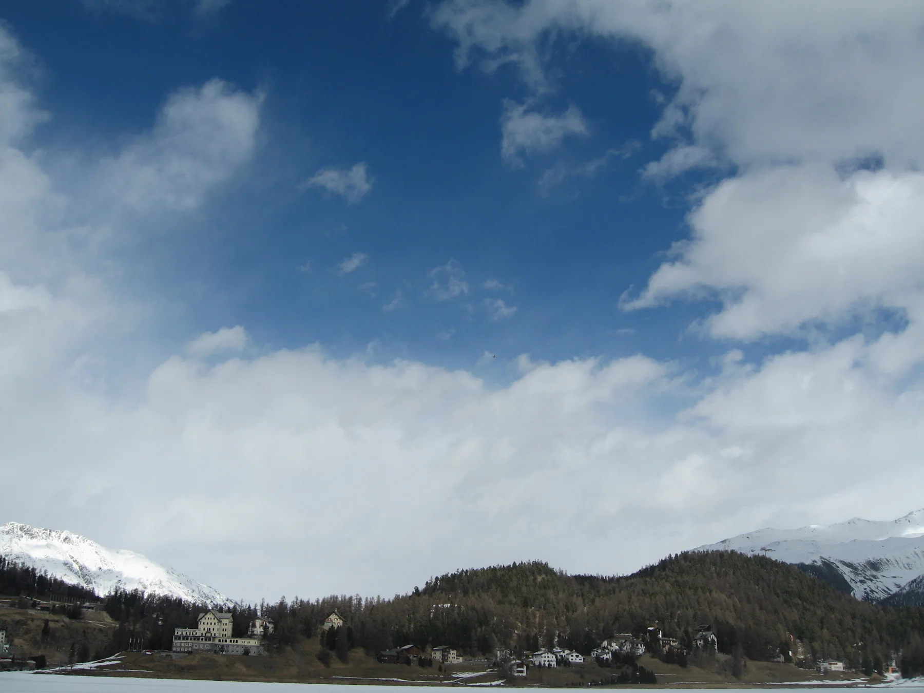 Large clouds cross a blue sky above a frozen lake and low wooded hills.
