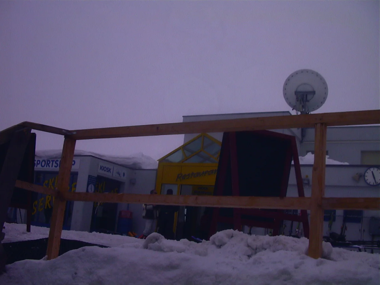 A wooden railing and terrace furnishings sit in snow beneath a pale, misty sky.