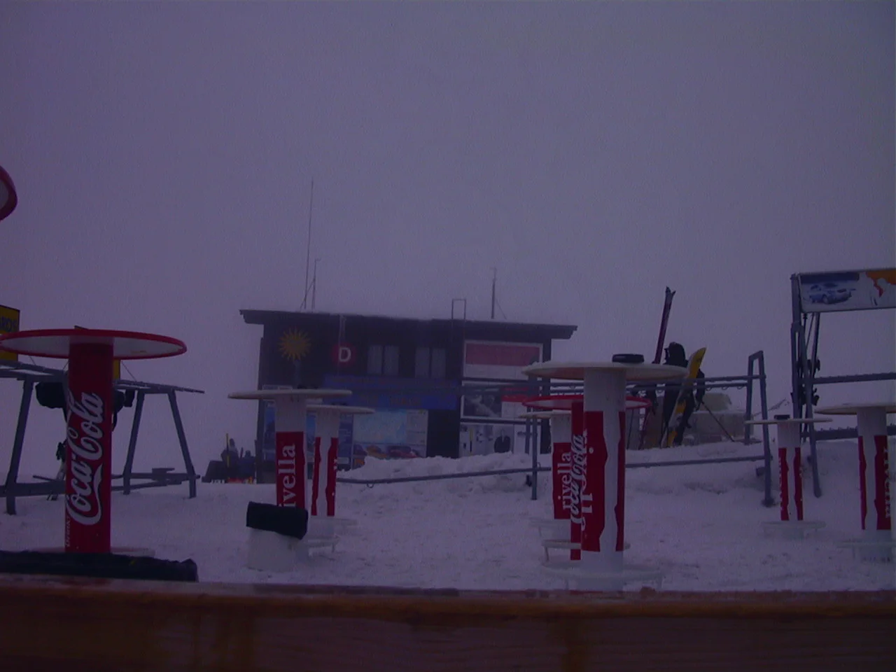Ski equipment, signs and a low building occupy a snowy lift area in muted light.