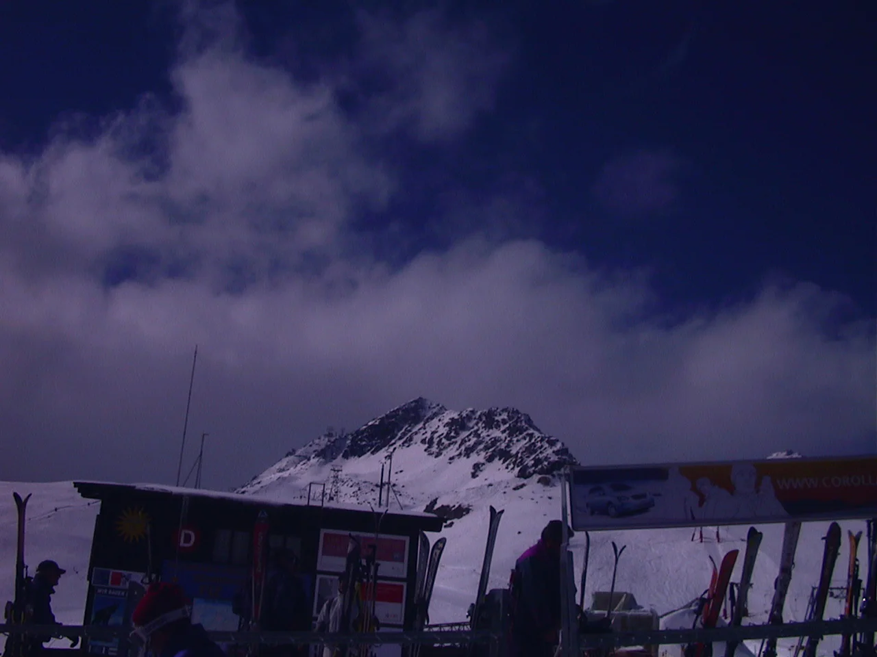 A rocky snowy peak rises behind a low ski-area building and drifting clouds.