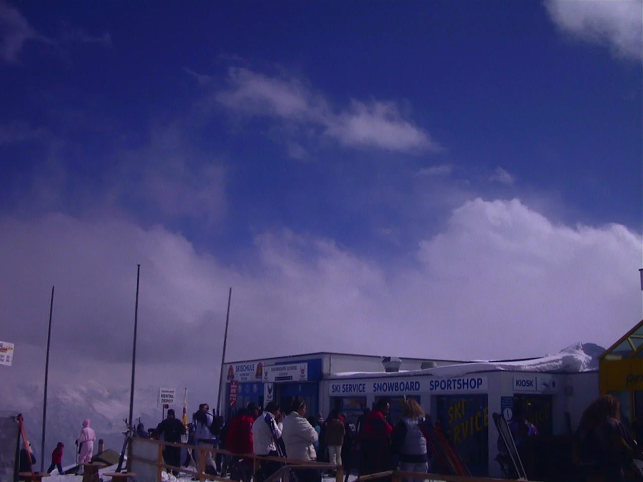 People gather near a mountain building beneath a blue opening in heavy clouds.